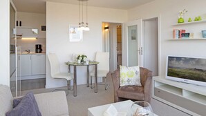 Una sala moderna con sofá, una mesa de centro con un libro y objetos decorativos, un televisor sobre un mueble bajo, una zona de comedor con sillas y una mesa, y una cocina al fondo.