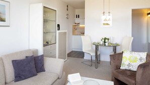 Una sala moderna con un sofá color beige, un sillón marrón y una mesa de centro de vidrio. Se ve una mesa de comedor con sillas y un área de cocina al fondo.