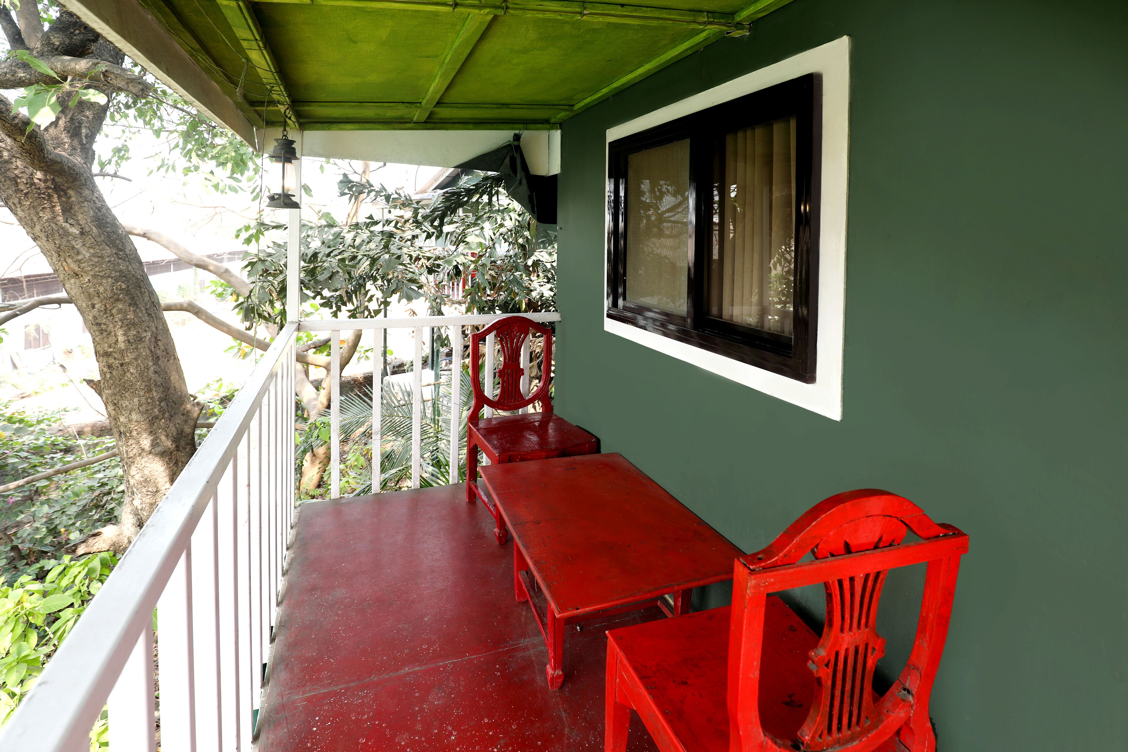 A covered balcony with a red table and chairs, a window with curtains, and a view of greenery.