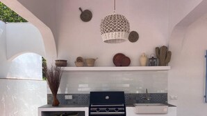 An outdoor kitchen with a white countertop, two wicker chairs, a hanging light fixture, and various decorative items on the wall.