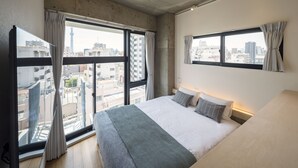 Una habitación de hotel moderna con una cama grande, dos ventanas con cortinas, vista a la ciudad y un techo de concreto.