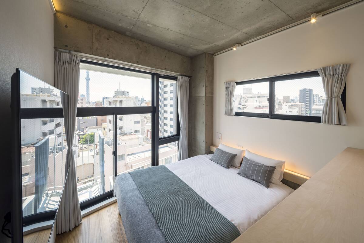 A modern hotel room with a large bed, two windows with curtains, a city view, and a concrete ceiling.