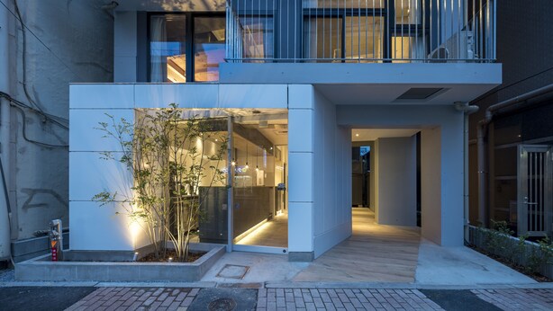 Un edificio moderno de dos pisos con entrada de vidrio, un pequeño jardín exterior y una escalera visible al nivel superior.