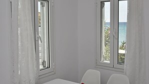 Ein heller Essbereich mit Tisch, Stühlen und einer Fruchtschale. Zwei Fenster mit Vorhängen, eines davon bietet Meerblick.