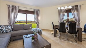 A modern living room with a grey sofa, a dining table with chairs, and large windows with curtains. - Renta Villa Georgios, with Pool, BBQ & Playground, Close to the beach (Perama)