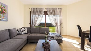 A modern living room with a grey sectional sofa, a coffee table with a vase and wine glasses, a dining table with chairs, and a large window with curtains. - Renta Villa Georgios, with Pool, BBQ & Playground, Close to the beach (Perama)