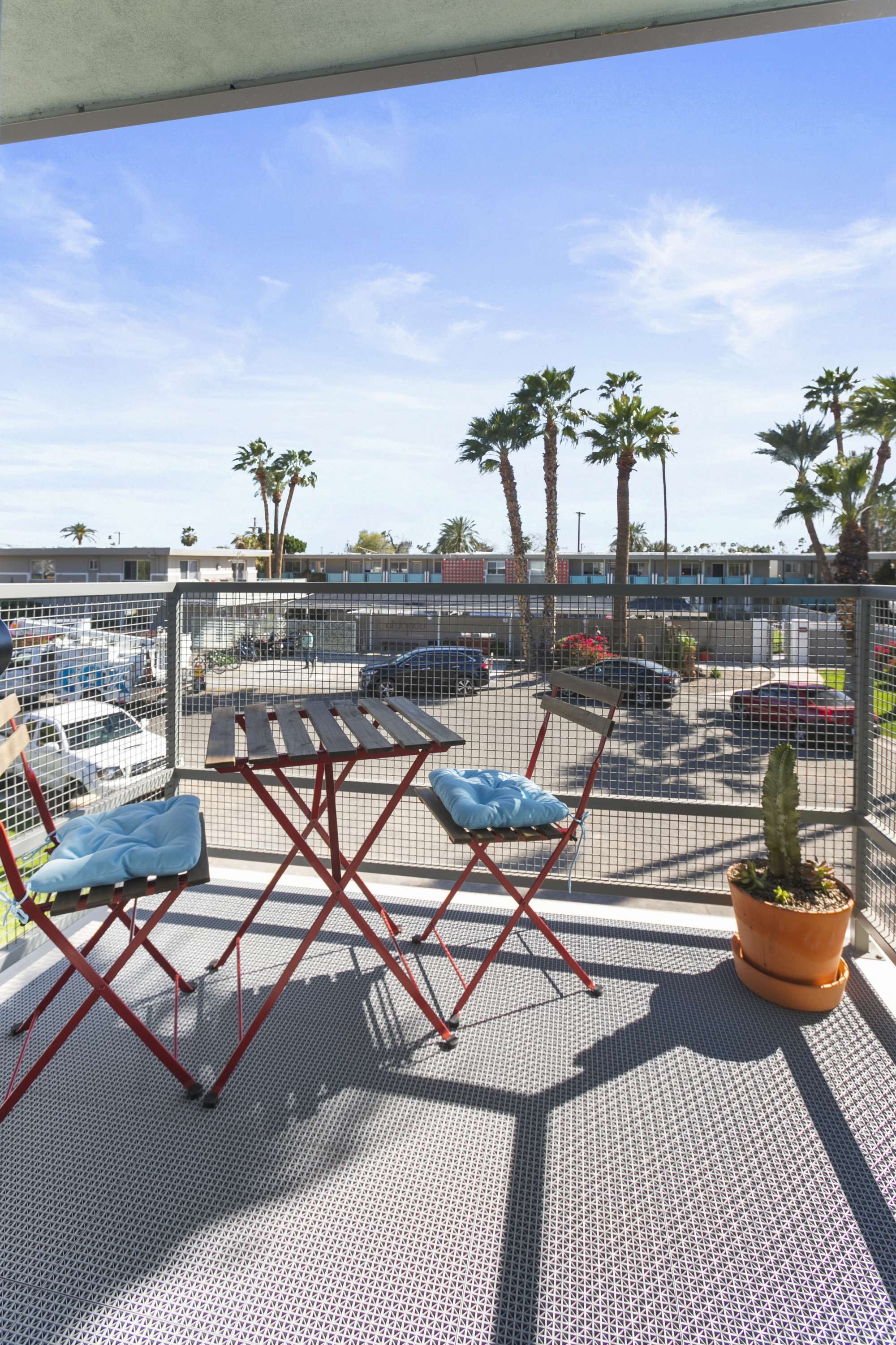 Terraço no telhado com mesa e cadeiras, um vaso de planta e vista para um estacionamento com carros e palmeiras.