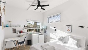 Une chambre d’hôtel compacte comprenant un coin cuisine, un lit avec du linge de maison blanc, une petite table à manger et un ventilateur de plafond.