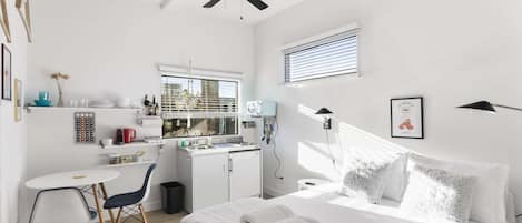 A compact hotel room with a kitchenette, a bed with white linens, a small dining table, and a ceiling fan.