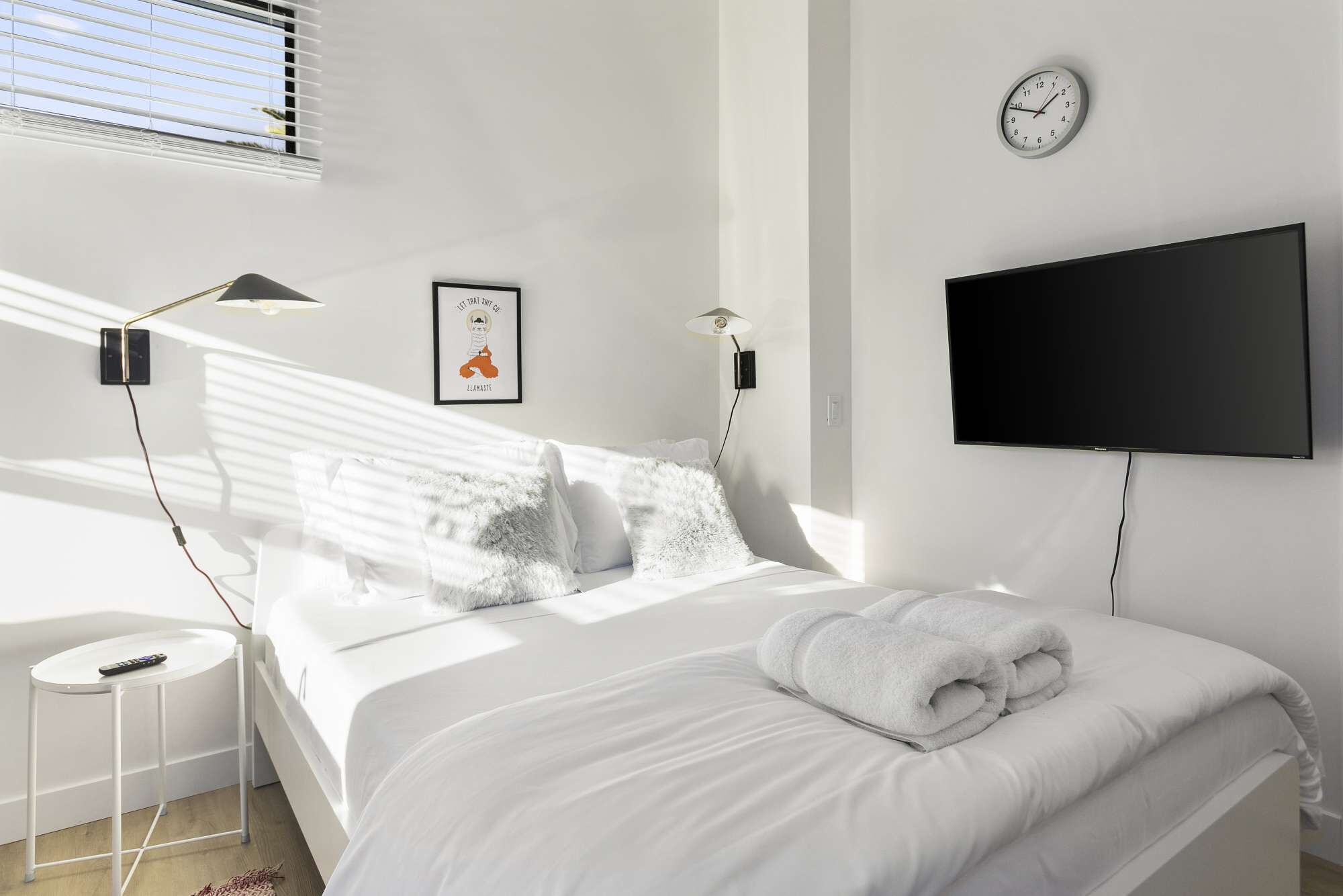 A modern bedroom with a bed, a bedside table with a remote, a wall-mounted TV, and a clock.