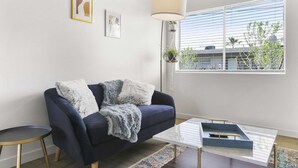 Ein modernes Wohnzimmer mit einem dunkelblauen Sofa, einer flauschigen weißen Decke und einem Glastisch. An der Wand befinden sich gerahmte Kunstwerke, eine hängende Pflanze und ein Blick ins Freie durch ein Fenster mit Jalousien.