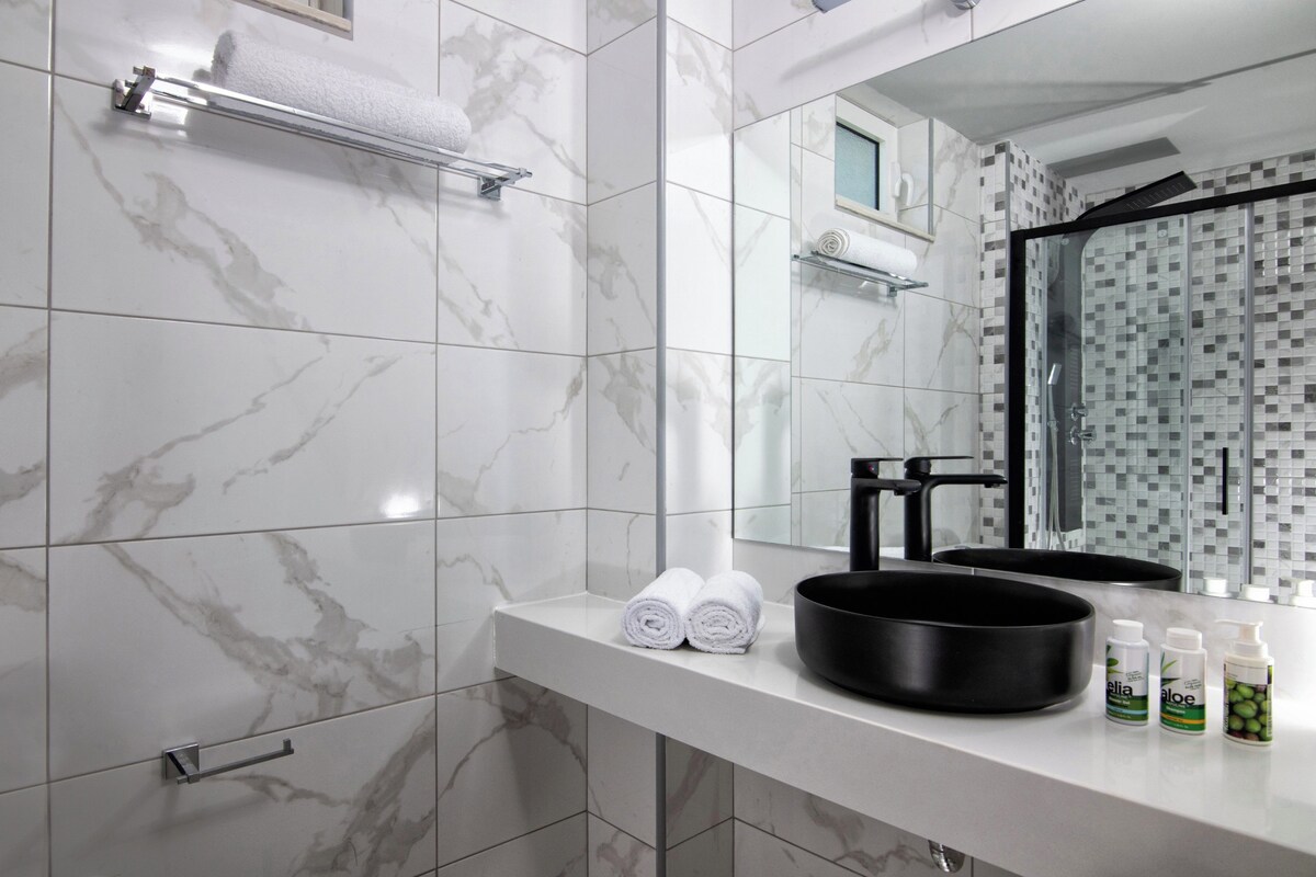 A bathroom with marble tiles, a black basin, rolled towels, and toiletries on the counter.