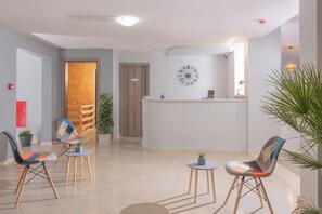 A reception area with a wooden reception desk, two chairs with colorful upholstery, a round wall clock, and a potted plant. - Golden Sunlight (Hersonissos)