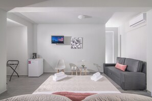 A modern hotel room with a bed, a gray sofa, a small table with a plant, a coffee machine, and a TV on the wall.