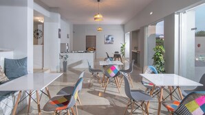 A modern dining area with white tables and chairs, a kitchen in the background, and large windows offering a view.
