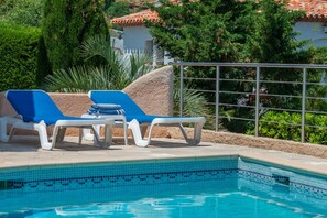 A pool area with two blue lounge chairs and a small table, surrounded by greenery and a tiled pool.