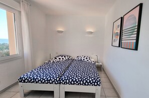 A small, neatly made bed with a polka dot bedspread in a room with white walls, a window with curtains, and two wall-mounted lights.