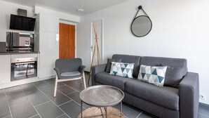 Un espacio de estar moderno con un sofá gris, dos cojines estampados, una mesita redonda, un televisor montado sobre la cocina y una puerta de madera.