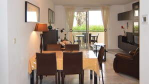 A dining area with a table and chairs, a sofa, a TV, and a balcony with curtains.