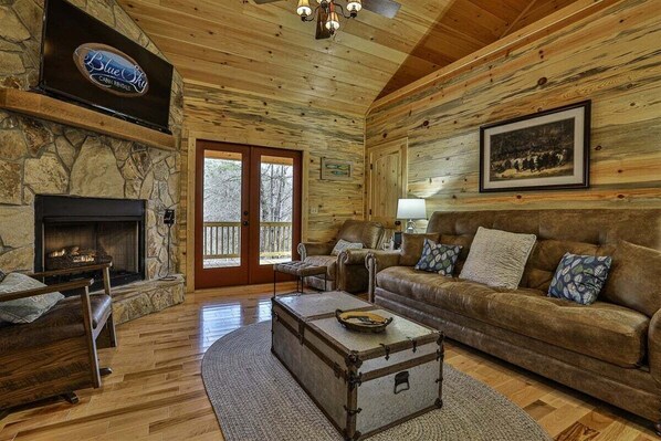 A log cabin interior with a stone fireplace, leather sofa, and a mounted television.