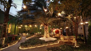 Een buitenplaats met een tuin, een centrale boom versierd met lichtjes, omringd door zitplaatsen en paden.