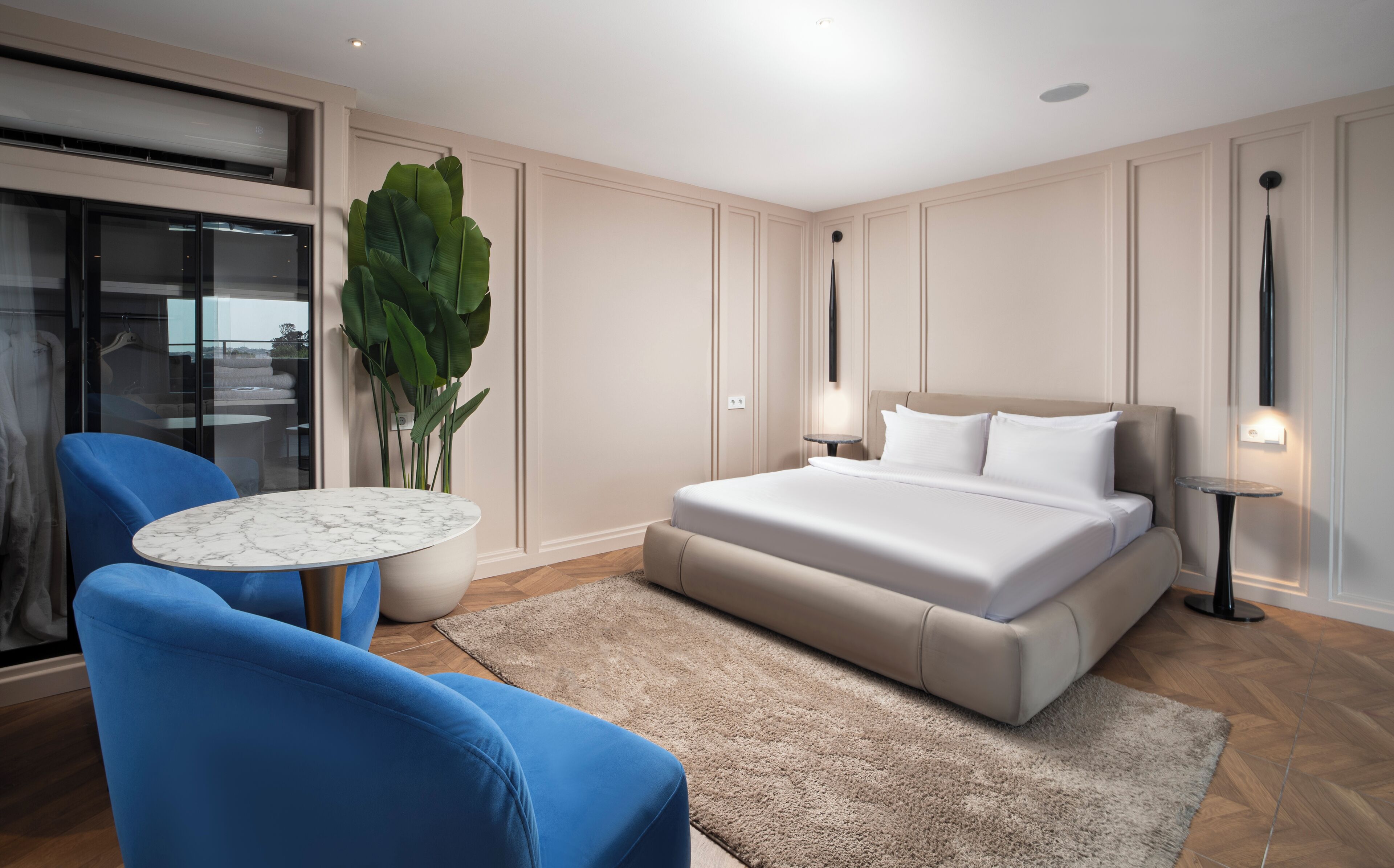 A modern hotel room with a large bed, a blue armchair, a small round table, and a large potted plant.