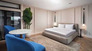 A modern hotel room with a large bed, a blue armchair, a small round table, and a large potted plant.