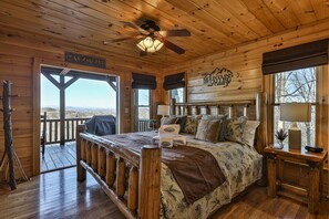 A wooden cabin bedroom with a bed, a ceiling fan, a window with a view, and a sign on the wall.