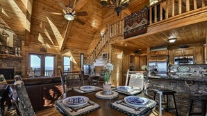 Interior de uma cabana de madeira com mesa de jantar, cadeiras, área de cozinha e varanda com vista.