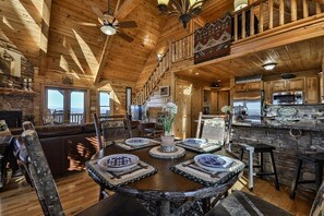 A wooden cabin interior with a dining table, chairs, a kitchen area, and a balcony with a view.