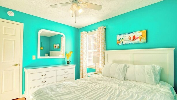 A bedroom with a bed, a white dresser, a ceiling fan, a window with curtains, and a painting on the wall. - Relax at the serene and spacious duplex nestled in the peaceful, family-friendly neighborhood. (Johnson City)