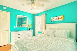 Une chambre à coucher avec un lit, une commode blanche, un ventilateur de plafond, une fenêtre avec des rideaux et un tableau accroché au mur.