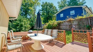 A wooden deck with a round table and chairs, a covered umbrella, and a blue house in the background. - Relax at the serene and spacious duplex nestled in the peaceful, family-friendly neighborhood. (Johnson City)
