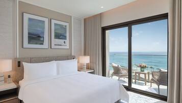 A hotel room with a large bed, two bedside tables with lamps, a view of the ocean through a sliding glass door, and two framed artworks on the wall.