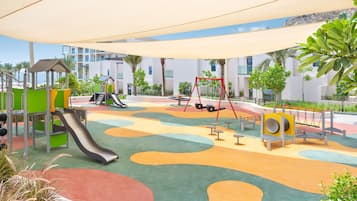 A playground with slides and swings, surrounded by palm trees and modern buildings.