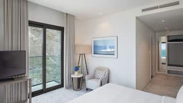 A hotel room with a large bed, a television on a stand, a small side table with a lamp and vase, a chair, a framed picture on the wall, and a view of the outdoors through a large window.