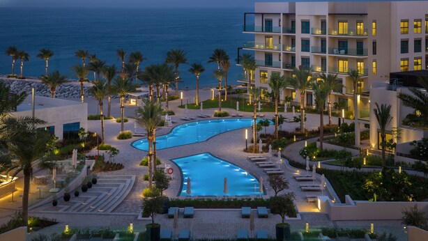 A resort pool area with a curved pool, palm trees, and a view of the ocean. - Address Beach Resort Fujairah (Al Aqah)