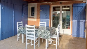 A covered outdoor dining area with a stone-tiled floor, a table with a patterned tablecloth, and white chairs. Blue doors and shutters are visible on the walls.