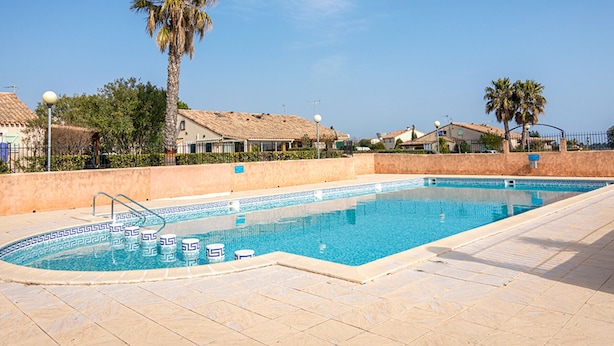 A swimming pool with a tiled deck, surrounded by palm trees and houses with tiled roofs.