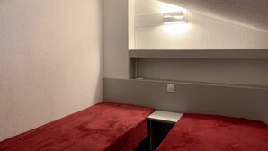 A small, narrow room with two beds covered in red blankets, a white wall, and a light fixture above the beds.