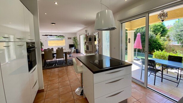 A modern kitchen with white cabinets, a black countertop, and a large island. There is a dining area with a table and chairs, and a patio with a table and chairs outside. - 3-bedroom Villa in Mèze with Garden and Air Conditioning (Mèze)