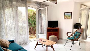 A living room with a sofa, a wooden coffee table, a wicker chair, a television on a wooden stand, and a view of greenery through the door. - Bright apartment in Bouzigues, 50m from beach, garden and parking (Bouzigues)