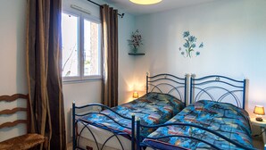 A bedroom with a metal bed frame, a wooden chair, a window with curtains, a wall-mounted shelf with flowers, and a wall decoration. - Air-conditioned house in Mèze with garden and private parking (Mèze)
