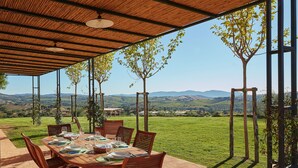 Uma área de refeições ao ar livre com uma mesa posta para quatro pessoas, rodeada por cadeiras de madeira, sob uma pérgula de madeira com vista para colinas ondulantes e um céu limpo.