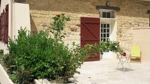 Un edificio in pietra con una porta rossa e persiane, un piccolo giardino con piante verdi e fiori rossi, e una sedia gialla in primo piano.