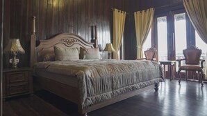 A large bed with a wooden headboard, two bedside tables with lamps, a chair, and a view of the outdoors through a window with curtains. - Asoka Resort Banten (Panimbang)