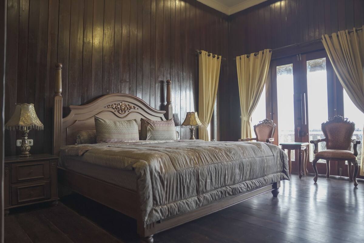 Un grand lit avec une tête de lit en bois, deux tables de chevet avec des lampes, une chaise et une vue sur l’extérieur par une fenêtre avec des rideaux.