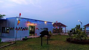 A building with a corrugated metal exterior, a covered outdoor seating area, and a small garden with a wooden bench. - Asoka Resort Banten (Panimbang)