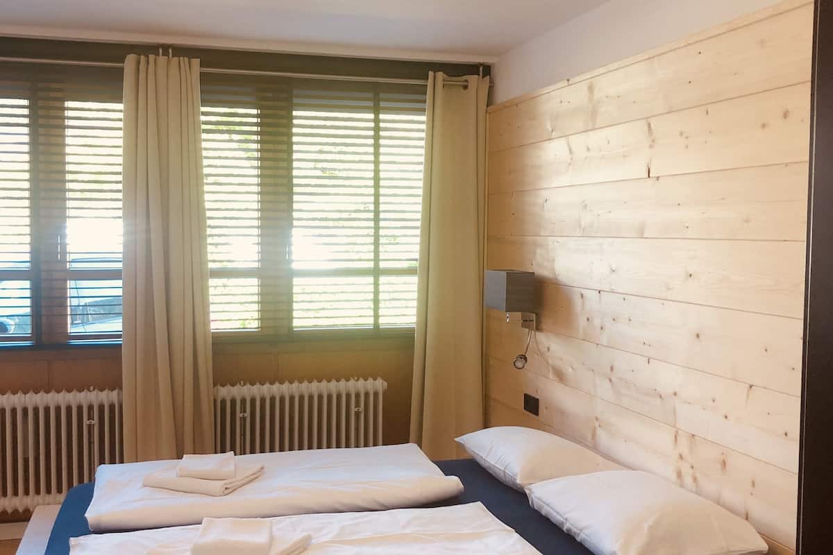 Una habitación con dos camas, un radiador, una pared de madera y una ventana con cortinas.