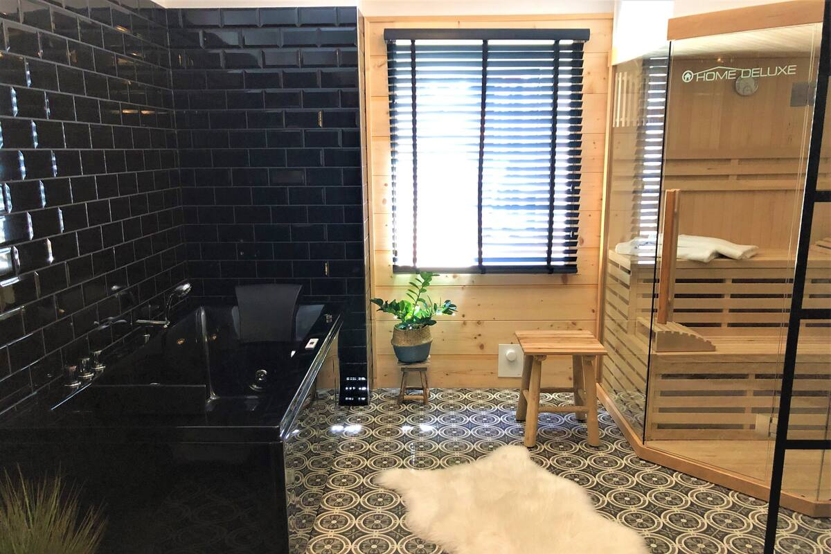 Un baño moderno con pared de azulejos negros, cabina de ducha con mampara de vidrio, banco de madera y planta en maceta.